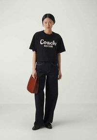 Black cropped t-shirt with "Coach New York" in white text, paired with dark blue wide-leg jeans. Holding a small brown handbag.
