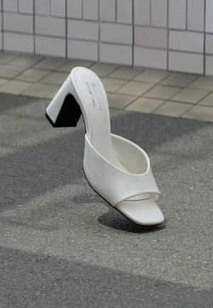 Singolo mule bianco a punta aperta con tacco a blocco, in piedi su asfalto grigio davanti a una parete piastrellata.