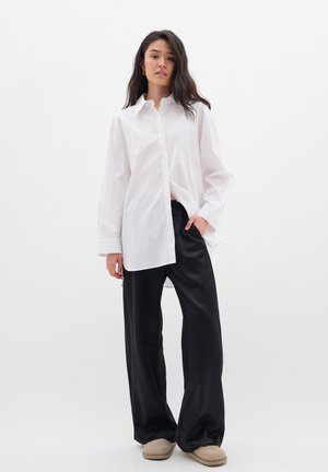 Camisa blanca de botones con mangas largas, combinada con pantalones anchos negros y zapatos tipo slip-on beige. Tejido suave con un ajuste relajado.