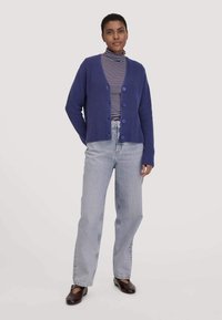 Frau steht und trägt eine blaue Strickjacke über einem gestreiften Rollkragenpullover, hellblaue Jeans und dunkelbraune flache Schuhe vor einem weißen Hintergrund.