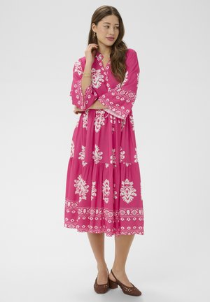Femme portant une robe midi rose à motifs floraux blancs, manches longues, et des chaussures à talons carrés marron, debout les bras croisés et regardant sur le côté.
