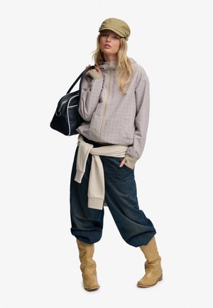 Ung kvinde iført beige kasket, ternet lynlåsjakke, løse mørke bukser, brune støvler og med en beige sweatshirt bundet om livet, der holder en sort håndtaske.