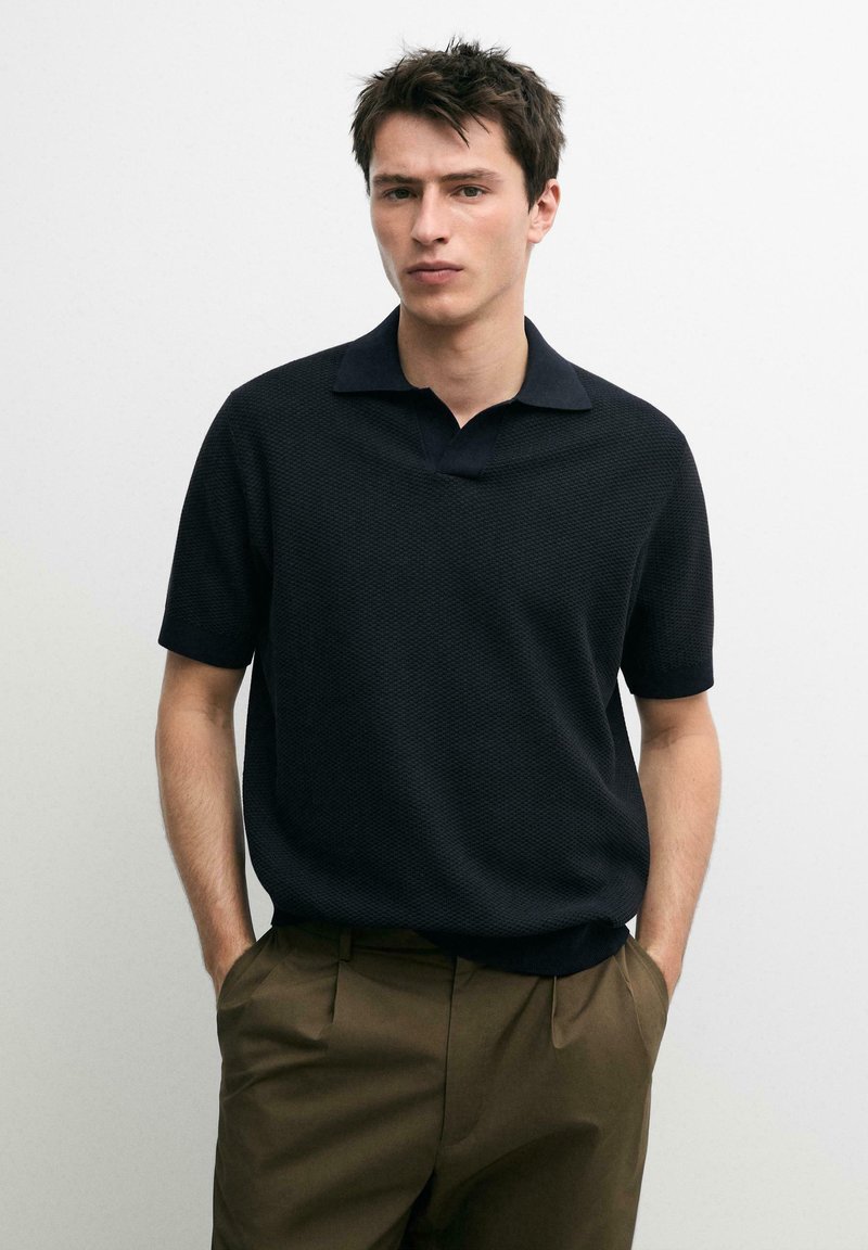 Giovane uomo che indossa una polo nera a maniche corte con texture e pantaloni verde oliva, in piedi con le mani nelle tasche contro uno sfondo bianco semplice.