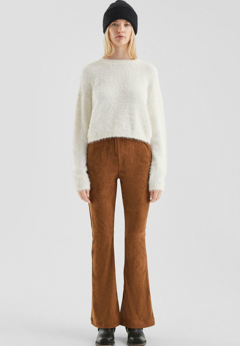 Vrouw in een zwarte gebreide beanie, witte fuzzy trui, bruine suède flared broek en zwarte laarzen, staand tegen een effen lichte achtergrond.