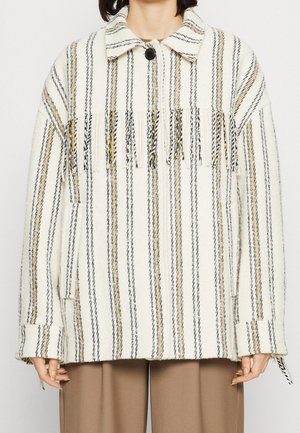 Veste rayée blanche et noire avec des accents jaunes, fermeture par boutons, large col et détails frangés au niveau de la poitrine. Matériau texturé.