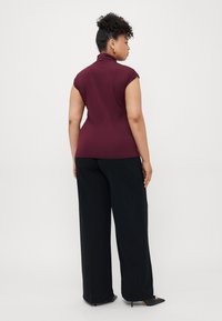 Kvinne med krøllete hår i en bun som har på seg gulløredobber, en ermeløs maroon topp, svarte vide bukser og svarte hæler, vendt bort.