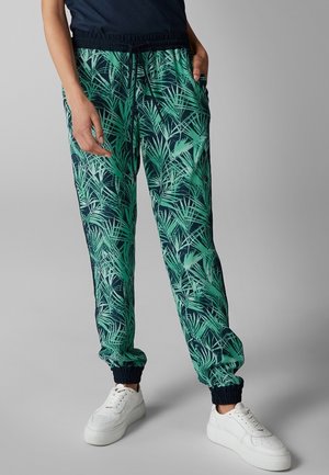 Persoon draagt groene en marineblauwe joggerbroek met tropisch bladmotief, elastische boorden en witte sneakers, staand tegen een effen grijze achtergrond.