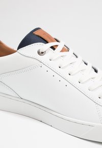 White sneaker with textured leather and a navy textile tongue. Features tan leather accents, round eyelets, and white laces.