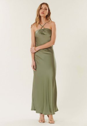 Jeune femme debout portant une robe longue en satin vert olive avec un col halter et des sandales dorées sur un fond clair uni.