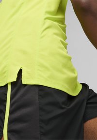 Top deportivo amarillo con una tela texturizada y transpirable; presenta una apertura lateral y se combina con unos pantalones cortos negros de acabado liso.