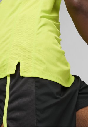 Żółta sportowa bluzka z teksturowanej, oddychającej tkaniny; posiada boczny rozcięcie i jest zestawiona z czarnymi szortami o gładkim wykończeniu.