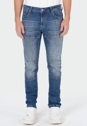 Homme portant un jean patchwork bleu ajusté et des baskets blanches, debout devant un fond clair uni.