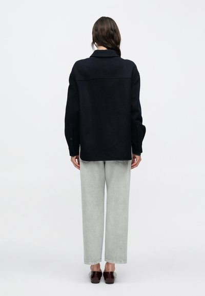 Μπλε ναυτικό πουκάμισο μεγάλης φόρμας με γιακά, μακριά μανίκια και μανσέτες με κουμπιά, συνδυασμένο με ανοιχτό γκρι τζιν κανονικής εφαρμογής και σκούρα παπούτσια τύπου slip-on.