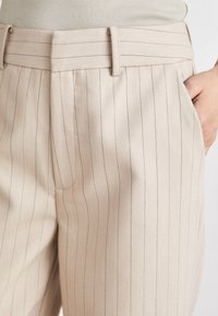 Pantalon beige à fines rayures avec ceinture assortie et poches latérales, porté avec un haut côtelé beige rentré, main dans la poche.