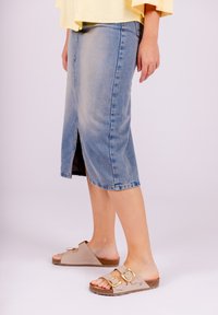 Denim midi skirt in light blue, featuring a front slit and frayed hem. Worn with beige slip-on sandals, accented by gold buckles.