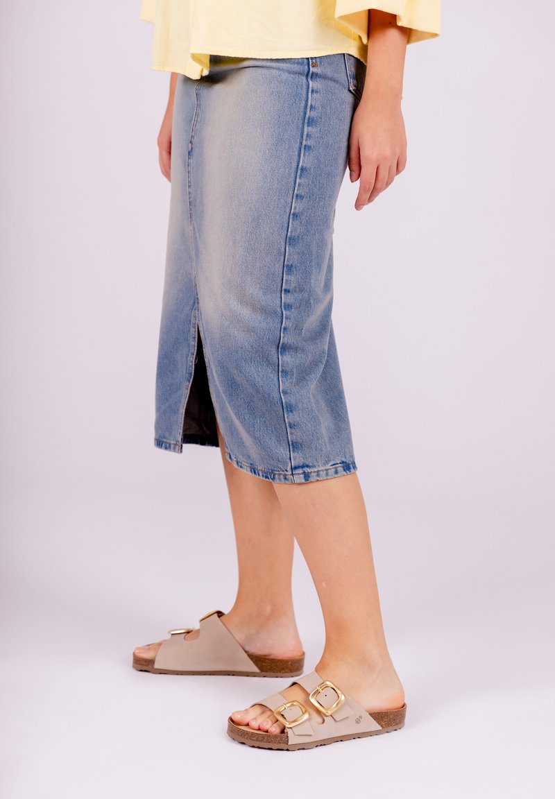 Denim midi skirt in light blue, featuring a front slit and frayed hem. Worn with beige slip-on sandals, accented by gold buckles.