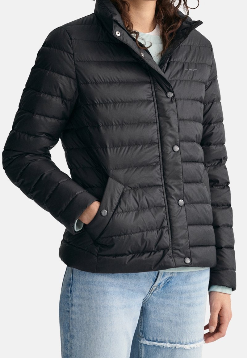 Black puffer jacket with a quilted design, snap buttons, side pockets, and a high collar. Worn over a light blue sweater.