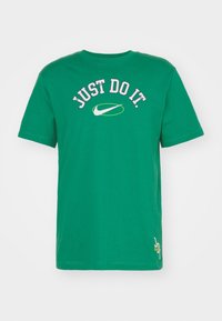Green cotton T-shirt with round neckline. Features "JUST DO IT" text in white and pink, and Nike logo in green. Short sleeves, relaxed fit.