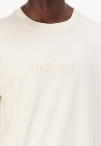 Gaišbeiges kokvilnas T-krekls ar apaļu kaklu un smalku izbrodētu "Hollister" logotipu priekšpusē. Gluda tekstūra, ikdienišķs dizains.