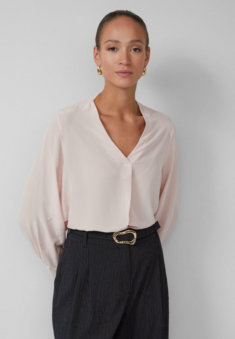Blouse rose clair à manches longues avec un col en V et une texture douce. Associée à un pantalon à rayures foncées et une boucle de ceinture ovale en or.