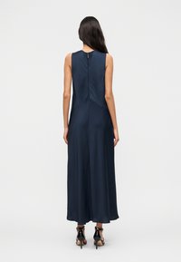 Γυναίκα με μακριά σκούρα μαλλιά, ντυμένη με αμάνικο μακρύ φόρεμα σε ναυτικό μπλε χρώμα και μαύρα σανδάλια με ψηλά τακούνια, όρθια πλάτη σε λευκό φόντο.