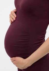 Graviditets Topp i vinröd, med en tight passform, ribbad textur och sidofoldningar, med en hand som vilar på en rundad mage.
