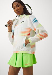 White windbreaker with colorful splashes in orange, green, and blue, paired with a neon green pleated skirt featuring a pocket and logo.