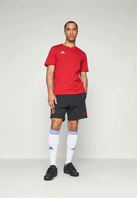 T-shirt vermelha da Adidas, calções pretos com o logótipo do Manchester United, meias brancas com riscas azuis, calçado preto, design casual e atlético.