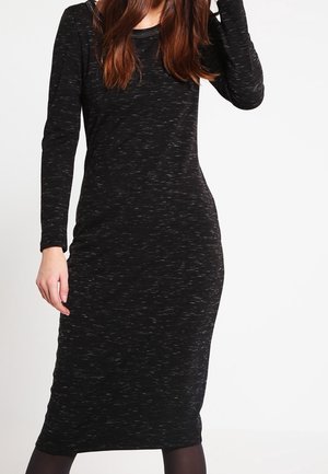 Frau trägt ein eng anliegendes, langärmliges schwarzes Midikleid mit dezenten weißen Sprenkeln und schwarze Strumpfhosen, steht vor weißem Hintergrund.