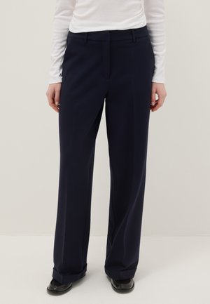 Femme portant un pantalon large bleu marine foncé et des chaussures noires, debout devant un fond clair uni, avec un haut blanc à manches longues.