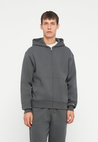Felpa grigia con zip, dotata di un ampio cappuccio, tasche frontali e polsini a costine. Presenta un piccolo logo ricamato sul petto. Tessuto in misto cotone.
