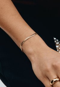 Złocona, teksturowana bransoletka o gładkim, zakrzywionym kształcie noszona na nadgarstku, cechująca się eleganckim, minimalistycznym wyglądem.