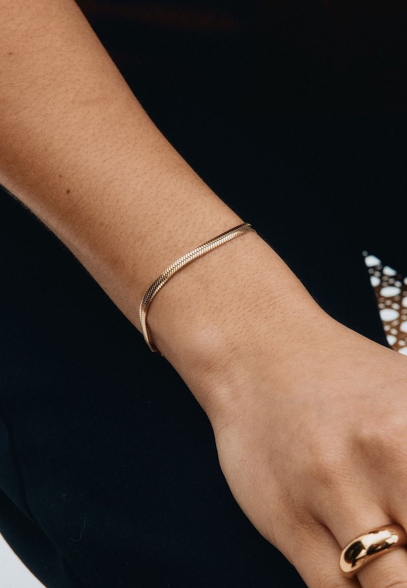 Złocona, teksturowana bransoletka o gładkim, zakrzywionym kształcie noszona na nadgarstku, cechująca się eleganckim, minimalistycznym wyglądem.