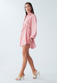 Rozā, ar garām piedurknēm un apkakli kleita ar pieguļošu vidukli un izplatu apakšu, izgatavota no mīksta auduma. Kombinēta ar gaismas krāsas augstpapēžu mule kurpēm ar atvērtām purngaliem.