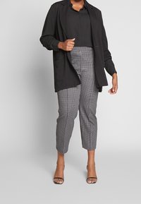 Blazer nero sopra una camicia nera, abbinato a pantaloni a quadri grigi e neri. Le scarpe hanno un design con cinturino. Tessuto morbido con una vestibilità su misura.