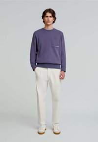 Paarse sweatshirt met een borstzak, gecombineerd met crèmekleurige geplooide broeken. Het model draagt witte sneakers met bruine zolen.