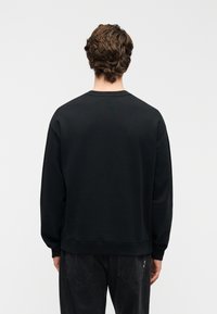 Μαύρο φούτερ με λαιμόκοψη crew, υφασμάτινη υφή, άνετη εφαρμογή, ριμπ μανσέτες και τελείωμα, χωρίς ορατά λογότυπα ή μοτίβα. Φορεμένο με σκούρα τζιν.