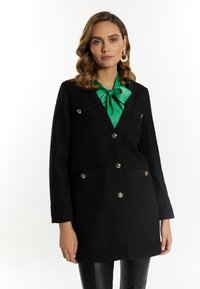 Cappotto nero con texture, dal design sagomato, bottoni dorati, due tasche frontali e un bordo a contrasto nero. Abbinato a un fiocco verde al collo.