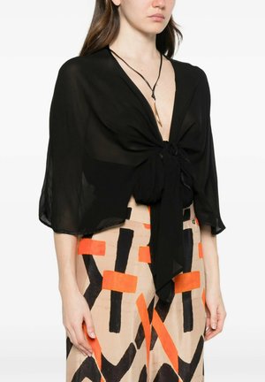 Donna che indossa una camicetta nera trasparente con nodo frontale e una gonna beige con motivi geometrici arancioni e neri audaci, più una collana in corda nera con ciondolo dorato.