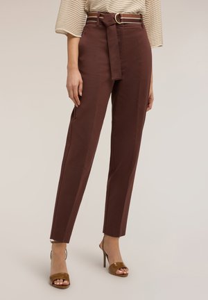 Donna che indossa pantaloni marroni a vita alta con taglio affusolato e cintura abbinata, e sandali marroni con tacco alto e punta aperta, in piedi su uno sfondo semplice.