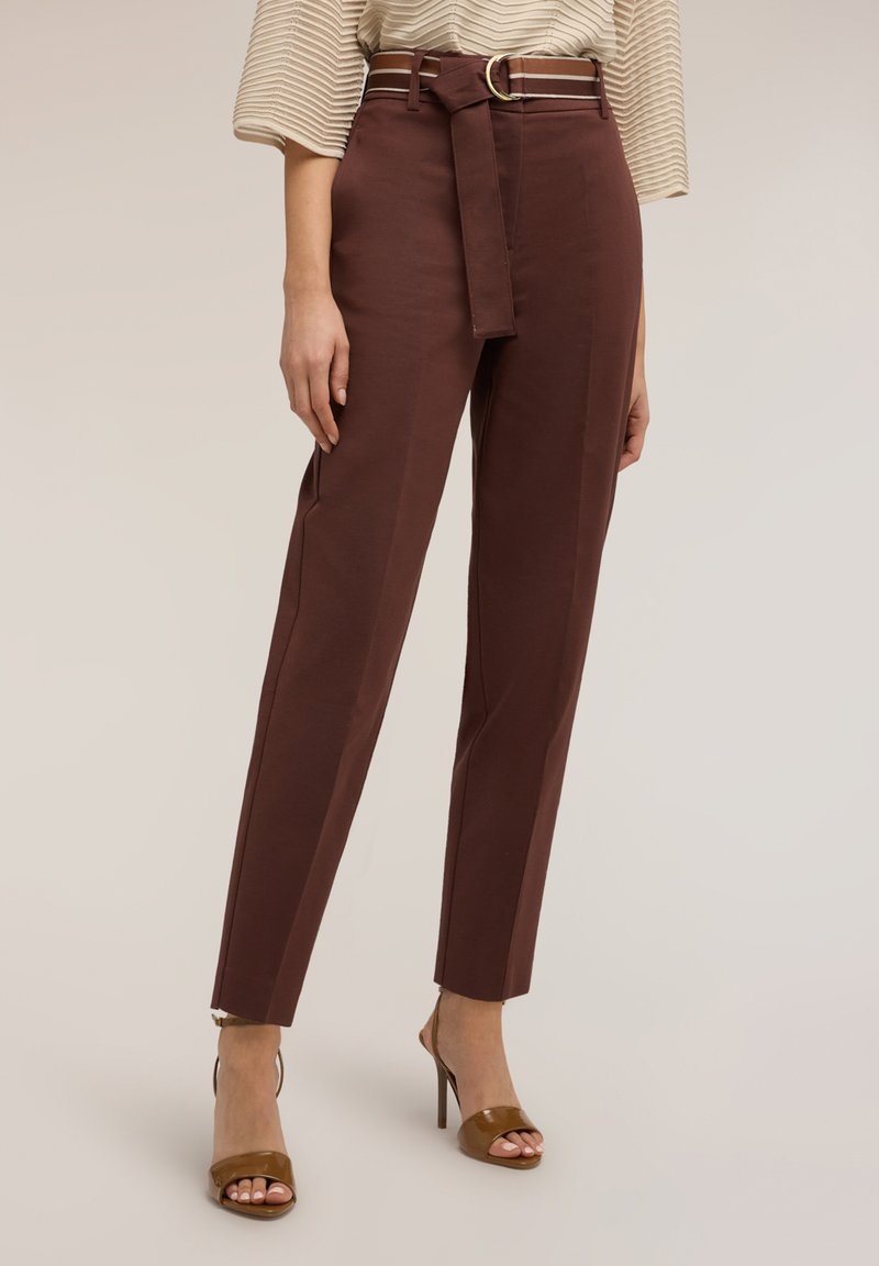 Frau trägt braune, hochgeschnittene, konisch zulaufende Hosen mit passendem Gürtel und braune Sandalen mit offenem Zehenbereich und hohen Absätzen, stehend vor einem schlichten Hintergrund.