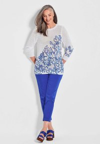 Weißer Netzpullover mit blauem Blumenmuster, kombiniert mit einfarbigen blauen Hosen. Das Modell trägt blaue Absatzsandalen.