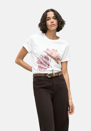 Vrouw met krullend haar draagt een wit T-shirt met roze bloemenprint, donkere broek en bruine riem, poseert met één hand achter de rug.