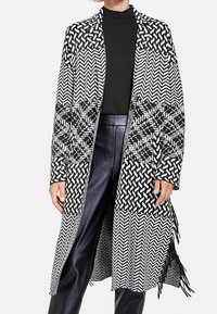 Svart och vit mönstrad långkappa med herringbone-design, fransig nederkant och öppen framsida, i kombination med en svart polo och läderbyxor.