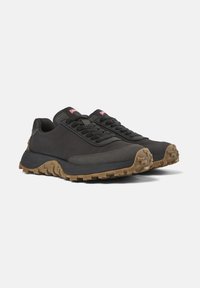 Sneakers neri a basso profilo con suole spesse marroni texture e lacci neri su uno sfondo bianco, posizionati affiancati a un angolo.