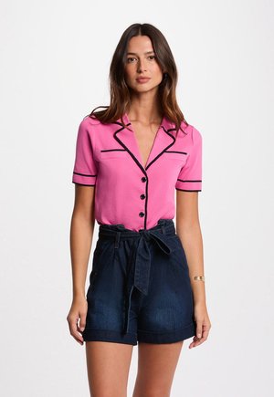 Model wearing a short-sleeved pink shirt with black trim and dark denim shorts, standing against a plain white background.