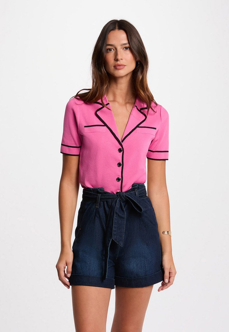 Model in een roze shirt met korte mouwen en zwarte biezen, en donkere spijkerbroekshorts, staand tegen een effen witte achtergrond.