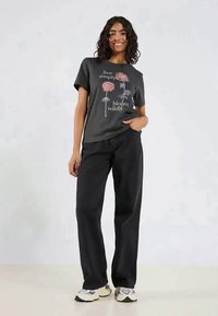Graues Baumwoll-T-Shirt mit floraler Grafikdruck in Rosa und weißen Text, kombiniert mit lockeren schwarzen Jeans und mehrfarbigen Turnschuhen.