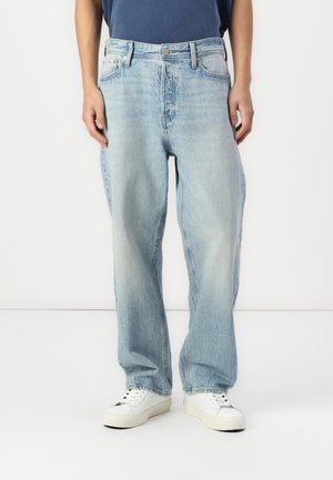 Osoba nosící volné světle modré džíny a bílé tenisky, stojící před jednoduchým bílým pozadím.