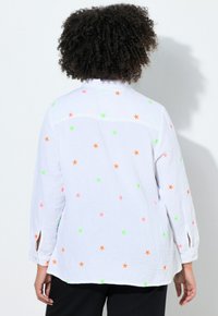 Person mit lockigem Haar, der ein weißes Langarmshirt mit neongelben, grünen und pinken Sternmustern trägt und vor einem schlichten weißen Hintergrund steht.